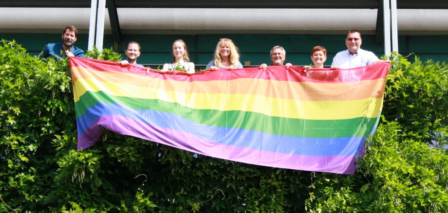 3 Frauen und 4 Männern stehen auf einem Balkon und halten eine Regenbogenflagge
