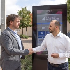 Digitales Gemeindeboard Handschlag vor dem digitalen Gemeindeboard zwischen zwei Männern (Dirk Koke und Bürgermeister Patrick Koch)