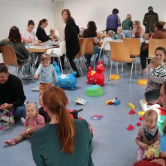 Einige Mütter mit ihren Kindern und ein Mann sitzen am Tisch und essen Kuchen. Weitere Mütter spielen mit ihren Kindern auf dem Boden