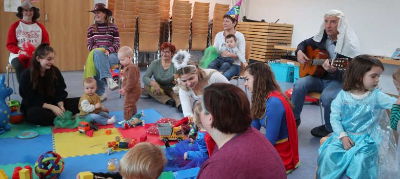 Verkleidete Kleinkinder sitzen mit Ihren Eltern auf dem Boden und spielen. Ein Mann spielt Gitarre