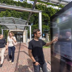 Zwei Frauen und ein Mann schauen sich das digitale Gemeindeboard an