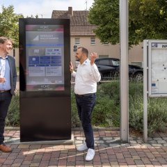 Zwei Männer (Dirk Koke und Bürgermeister Patrick Koch) reden vor dem neuen digitalen Gemeindeboard