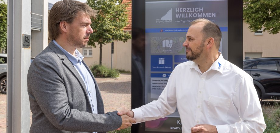 Handschlag vor dem digitalen Gemeindeboard zwischen zwei Männern (Dirk Koke und Bürgermeister Patrick Koch)