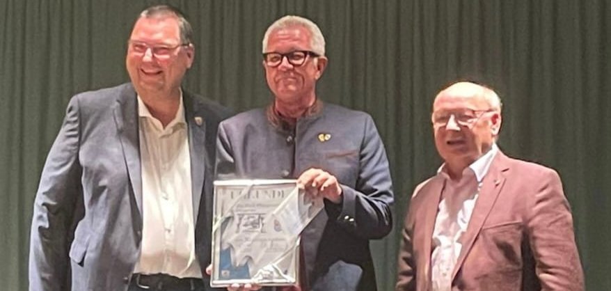 Drei Männer stehen auf einer Bühne. Der mittlere Mann hält eine Urkunde in der Hand.