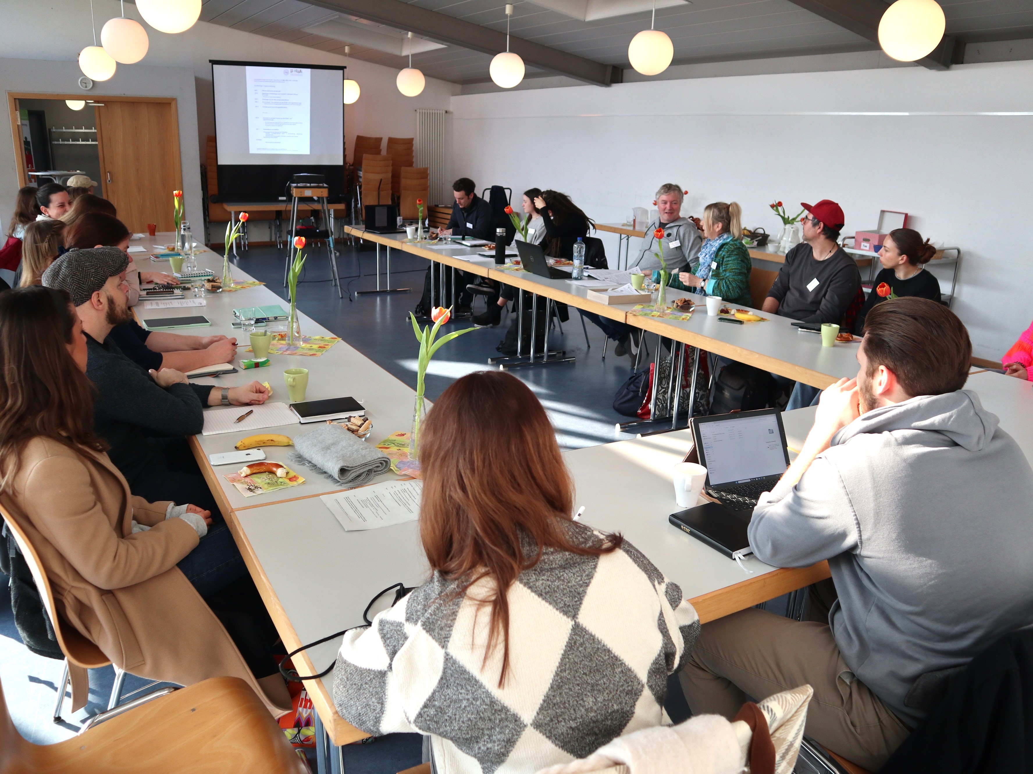 Menschen sitzen an einem u-förmigen Tisch und schauen auf eine Leinwand