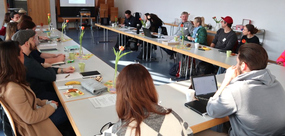 Menschen sitzen an einem u-förmigen Tisch und schauen auf eine Leinwand