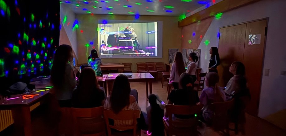 Mädchen schauen einen Film auf einer Leinwand. Der Raum ist dunkel und es leuchten bunte Lichter