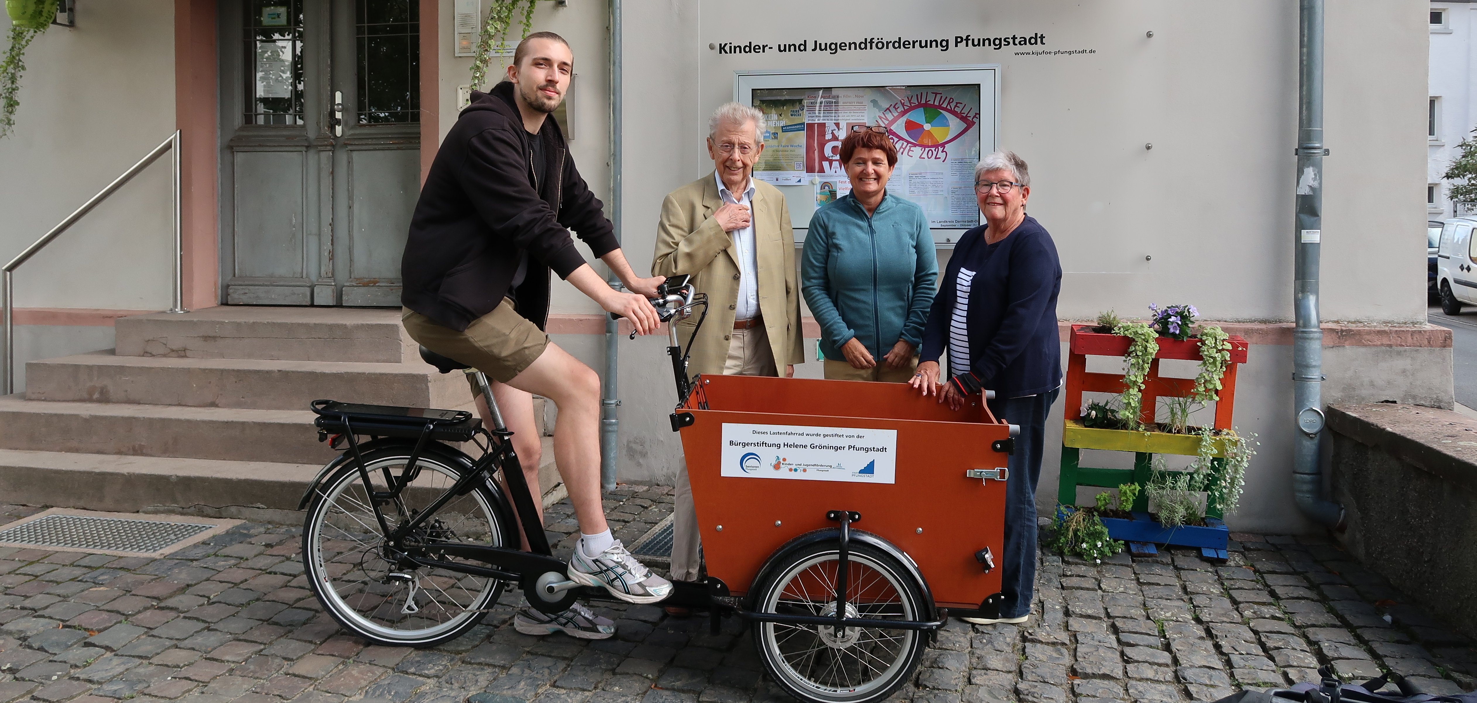 Ein Mann sitzt auf einem Lastenfahrrad, dahinter stehen zwei Damen und ein weiterer Mann