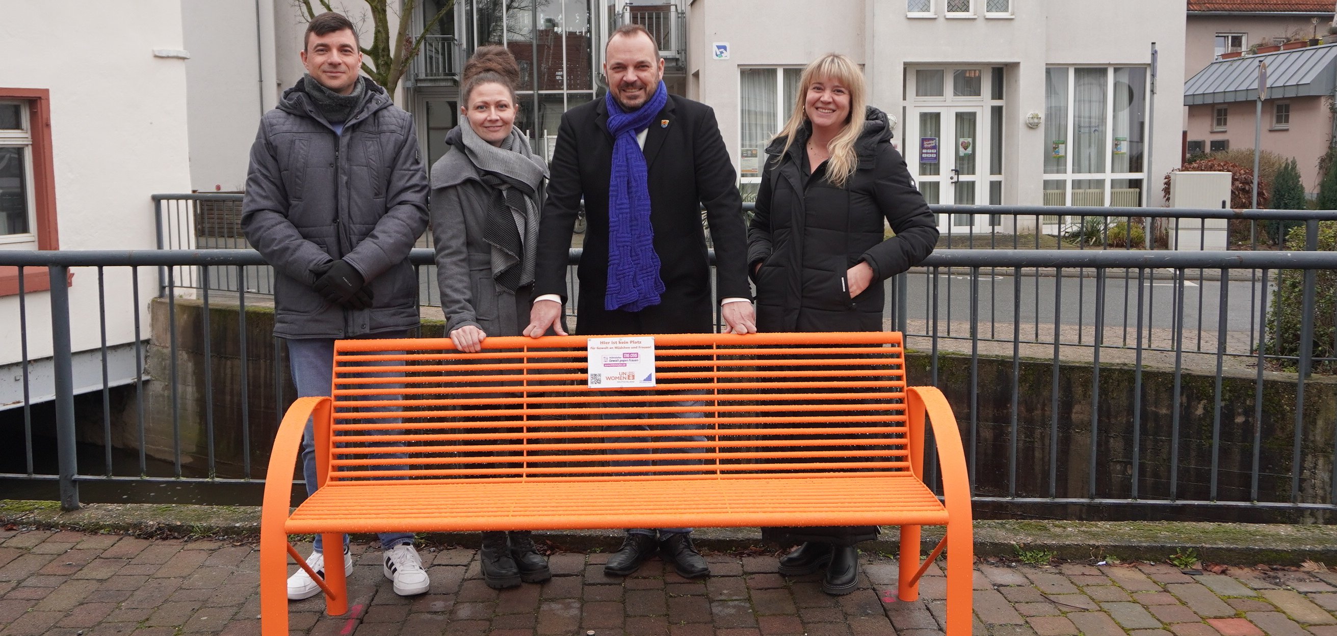 Zwei Frauen und zwei Männer stehen hinter einen orangefarbenen Parkbank