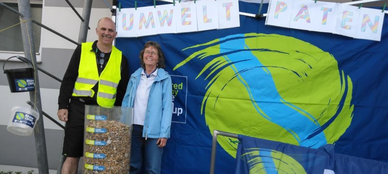 Eine Frau und ein  Mann stehen hinter einem durchsichtigen Behälter voller Zigarettenkippen