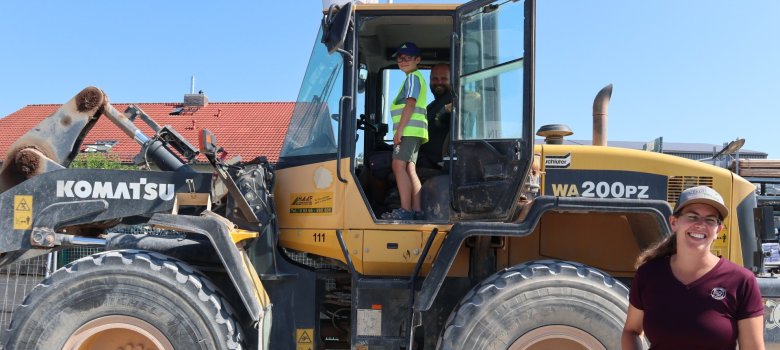 Im Vordergrund ist eine lächelnde Mitarbeitern des Natursteinzentrum Rhein-Main zusehen. Dahinter eine Baumaschine in dessen geöffneter Tür ein Junge mit Warnweste steht.