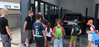 Kinder stehen neben zwei Mitarbeitern vor einer Garage und sehen sich ein darin geparktes Auto an.