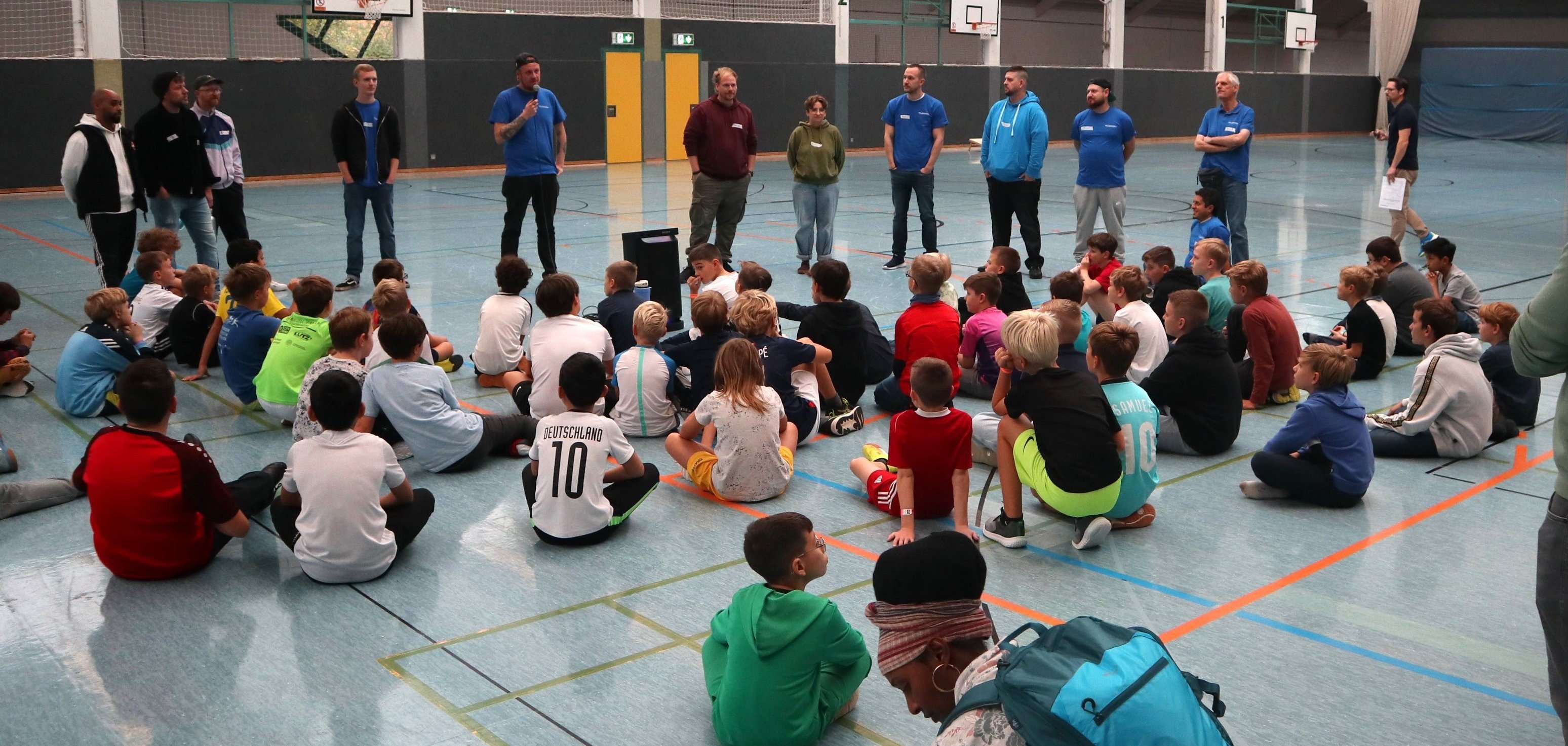Viele Kinder sitzen in einer Halle auf dem Boden und hören Erwachsenen zu, die ihnen etwas erzählen