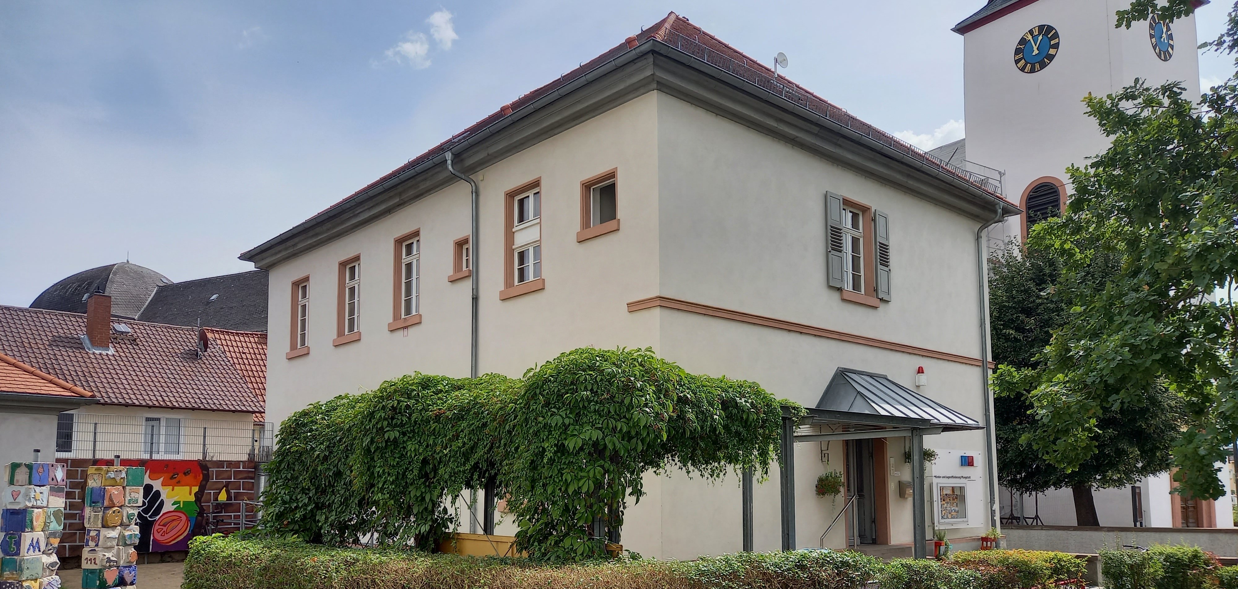 Weißes Gebäude mit Büschen im Vordergrund und einer Kirch im Hintergrund