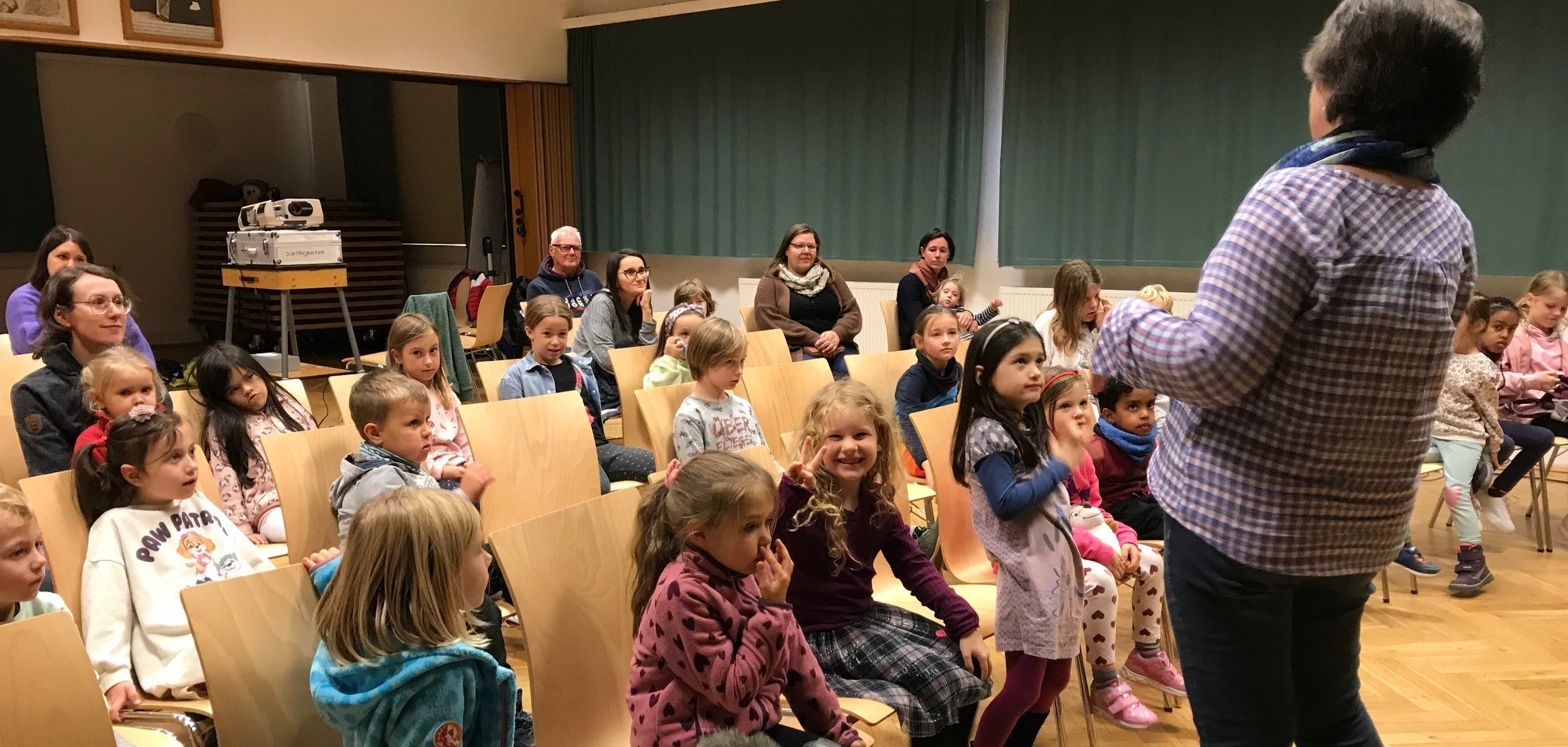 Eine Frau steht vor einer großen, auf Stühlen sitzenden Kindergruppe und hält einen Vortrag 