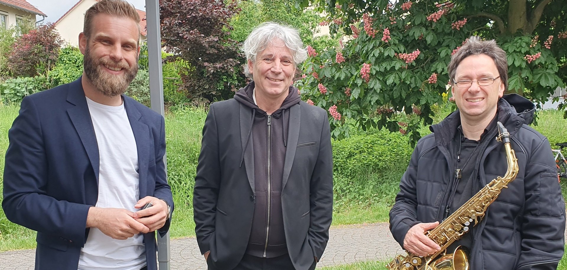 Drei Herren stehen unter einem Pavillon, einer hat ein Saxophon in der Hand