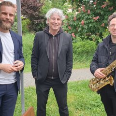 Zahlreiche Besucherinnen und Besucher genießen stimmungsvollen Abend mit Wein, Kulinarik und Musik im Park der Villa Büchner Drei Herren stehen unter einem Pavillon, einer hat ein Saxophon in der Hand