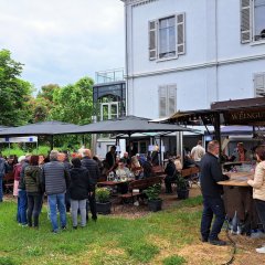 Zahlreiche Besucherinnen und Besucher genießen stimmungsvollen Abend mit Wein, Kulinarik und Musik im Park der Villa Büchner Menschen sitzen in einem Park unter schwarzen Sonnenschirmen und genießen Wein