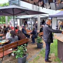 Zahlreiche Besucherinnen und Besucher genießen stimmungsvollen Abend mit Wein, Kulinarik und Musik im Park der Villa Büchner Menschen sitzen in einem Park unter Sonnenschirmen und genießen Wein. Ein Mann kauf Wein an einem Weinstand.