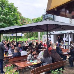 Zahlreiche Besucherinnen und Besucher genießen stimmungsvollen Abend mit Wein, Kulinarik und Musik im Park der Villa Büchner Menschen sitzen in einem Park unter Sonnenschirmen und genießen Wein